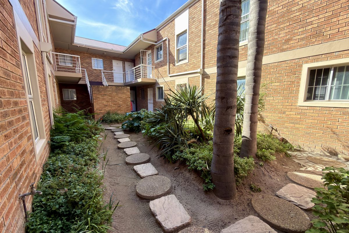 A view of the apartment complex courtyard featuring a brick facade, landscaped garden beds, and a stone pathway leading to the building entrance.