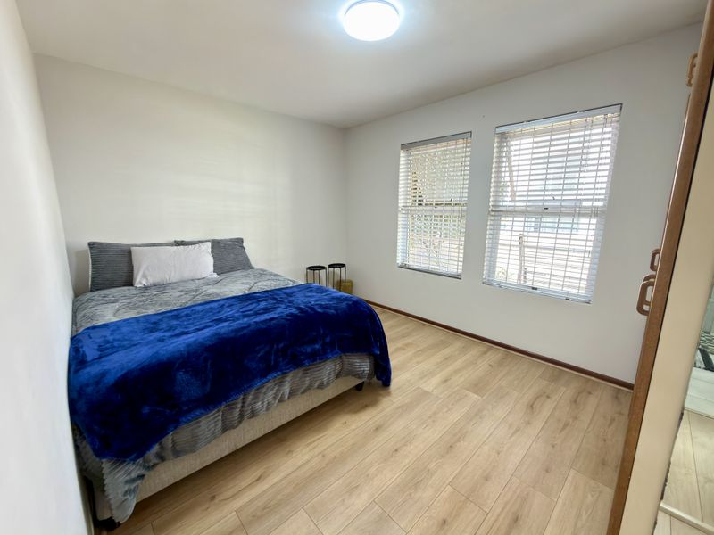 A well-lit bedroom featuring light-toned laminate flooring, dual windows with venetian blinds, and a built-in wardrobe.