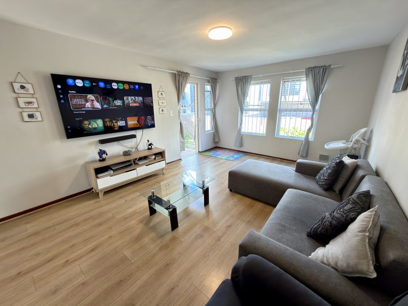 A well-maintained living room featuring light wood-look laminate flooring, large windows with security bars, and direct access to an outdoor area.