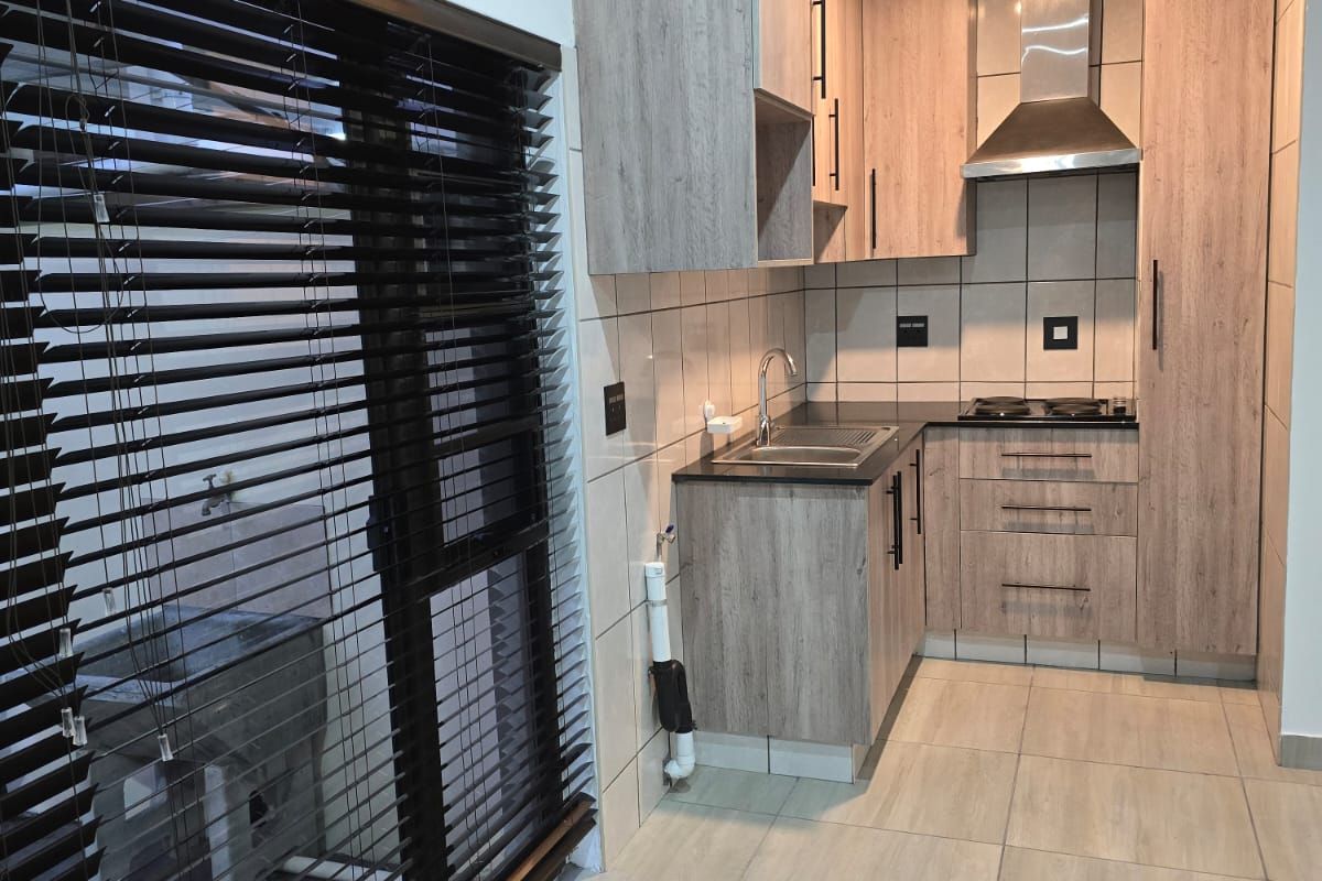 A modern, compact kitchen featuring light wood-grain cabinetry, a stainless steel extractor fan, a built-in hob, and neutral floor tiles.