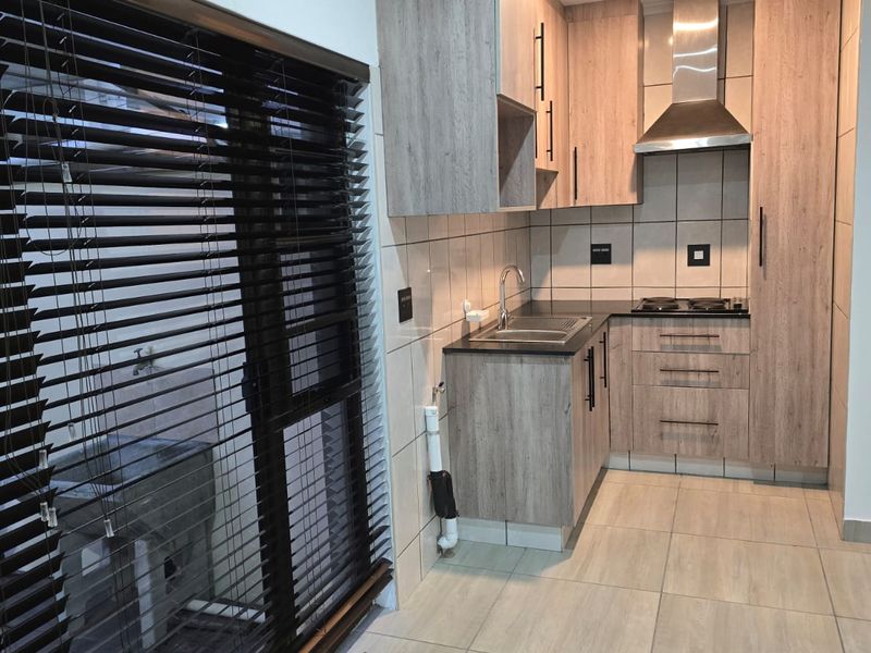 A modern, compact kitchen featuring light wood-grain cabinetry, a stainless steel extractor fan, a built-in hob, and neutral floor tiles.