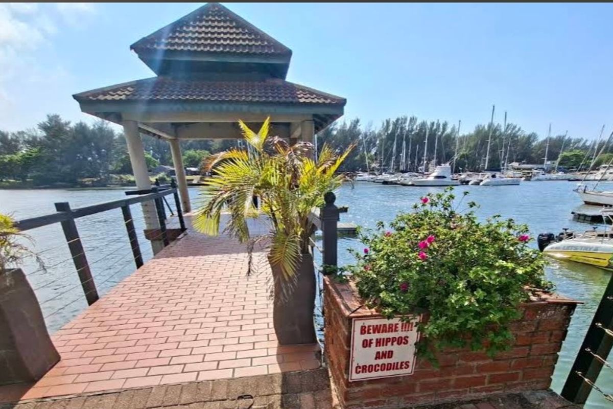 A waterfront jetty featuring a thatched gazebo, brick paving, and a brick planter box overlooking a marina with docked boats.