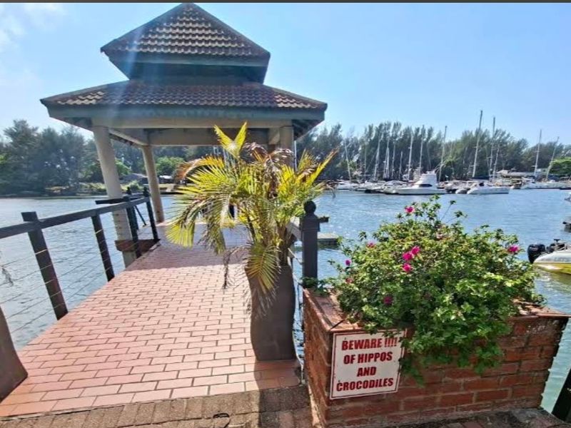 A waterfront jetty featuring a thatched gazebo, brick paving, and a brick planter box overlooking a marina with docked boats.