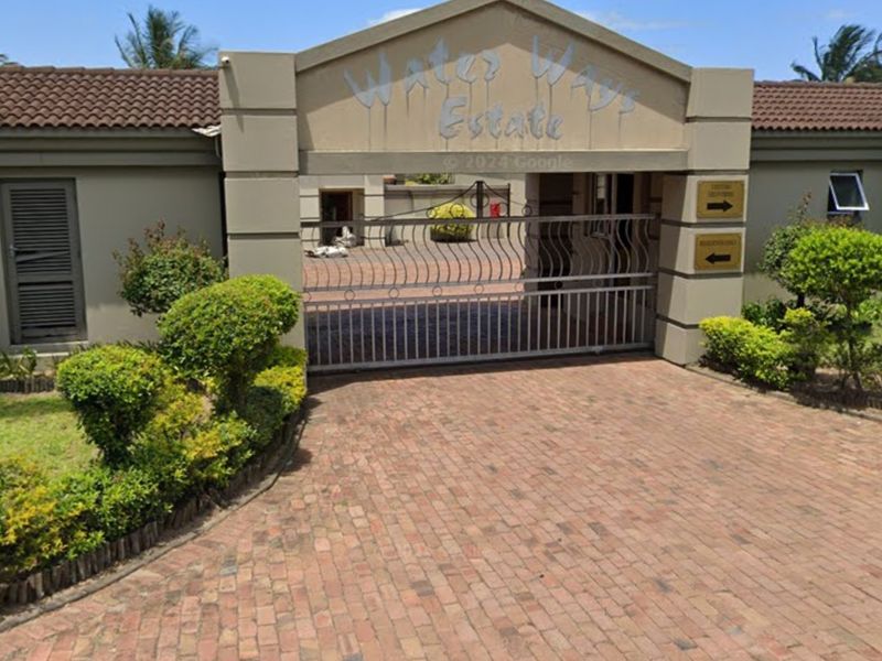 The gated entrance to Water Ways Estate featuring a brick paved driveway, secure access gate, and well-maintained garden landscaping.