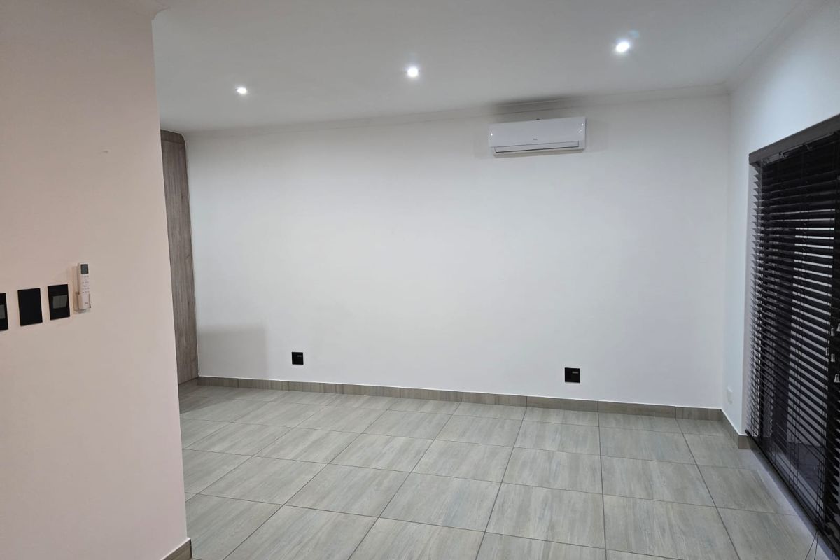 A bright, empty living area featuring modern tiled flooring, recessed downlights, and a wall-mounted air conditioning unit with dark venetian blinds.
