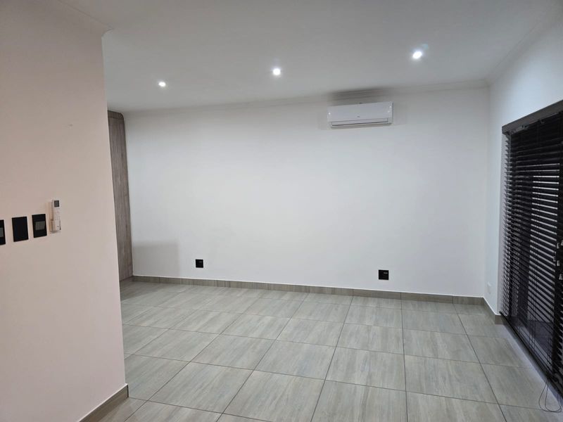 A bright, empty living area featuring modern tiled flooring, recessed downlights, and a wall-mounted air conditioning unit with dark venetian blinds.