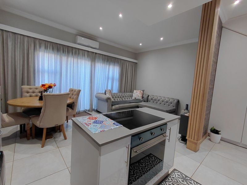 Modern open-plan living and dining area featuring a central kitchen island with integrated cooktop and oven, complemented by neutral tiled flooring and recessed lighting.