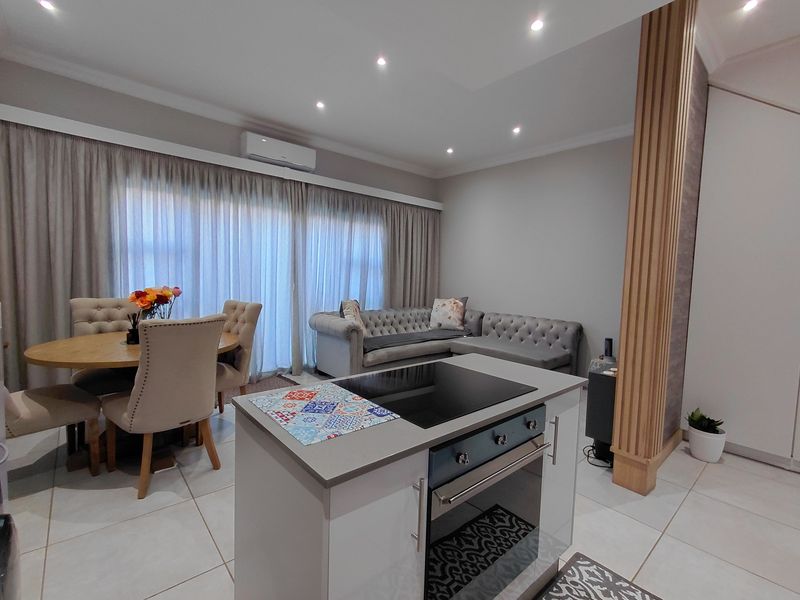 Modern open-plan living and dining area featuring a central kitchen island with built-in oven and cooktop, complemented by neutral tiled flooring and recessed lighting.