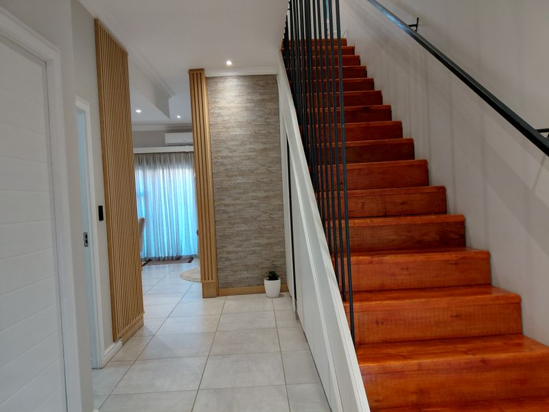 A bright, modern hallway featuring a polished timber staircase with a sleek metal balustrade, tiled flooring, and contemporary timber slat wall accents.