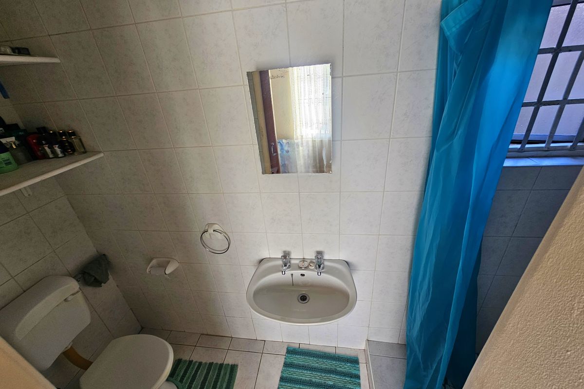 A compact bathroom featuring a wall-mounted basin, toilet, and shower area with tiled finishes and a window with security bars.