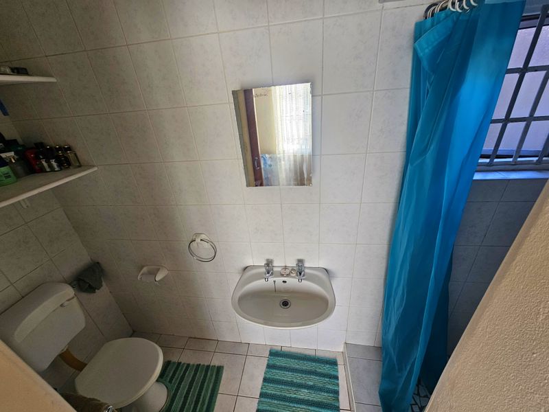 A compact bathroom featuring a wall-mounted basin, toilet, and shower area with tiled finishes and a window with security bars.