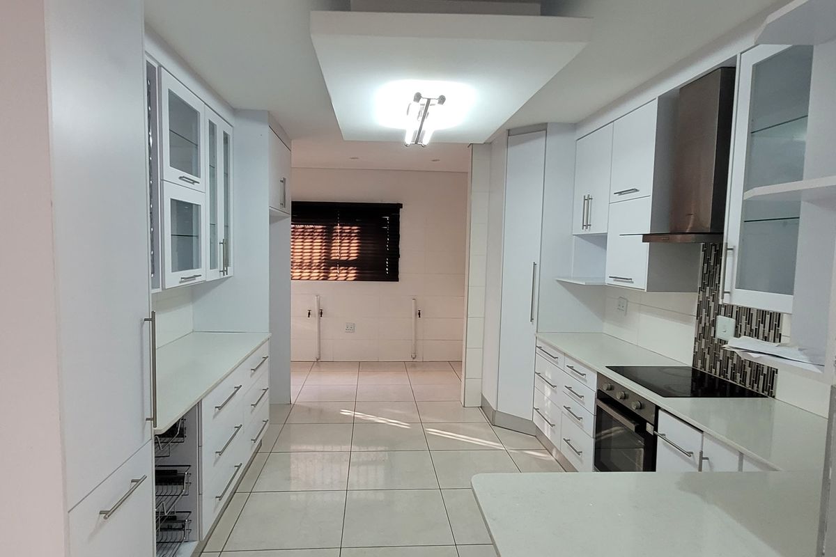 A modern, clean-lined kitchen featuring white cabinetry, stone countertops, integrated appliances, and tiled flooring, leading into an adjacent open-plan living area.