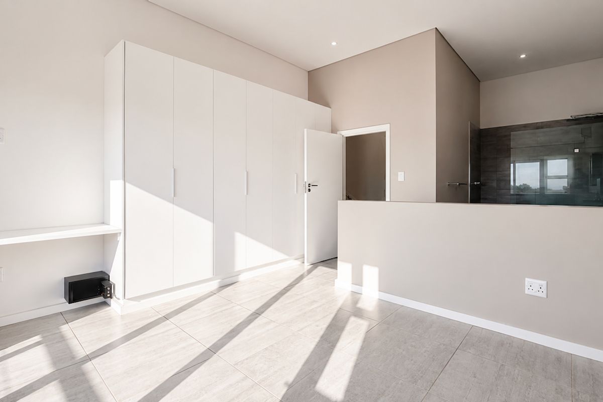 Modern, bright bedroom featuring built-in cupboards, a wall-mounted desk, and an open-plan en-suite bathroom with a glass shower enclosure.