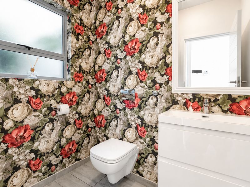 A modern bathroom featuring a wall-mounted toilet, white vanity with basin, and bold floral wallpaper, complemented by tiled flooring and a framed mirror.