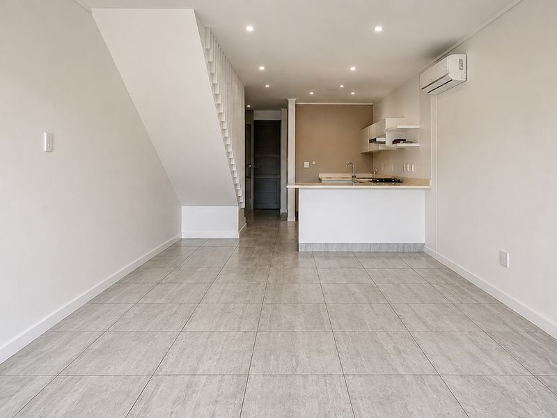 An open-plan living and kitchen area featuring neutral tiled flooring, a modern staircase, and a compact kitchen with built-in cabinetry and appliances.