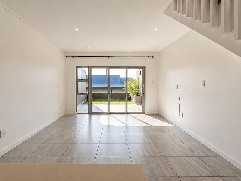 A bright, open-plan living area featuring neutral tiled flooring, recessed lighting, and large sliding glass doors that provide access to an outdoor space.