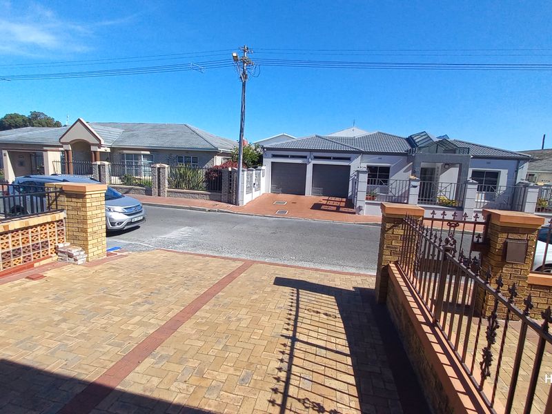 A street-level view of the property frontage, featuring a paved driveway, brick boundary walls with wrought iron fencing, and a double garage.