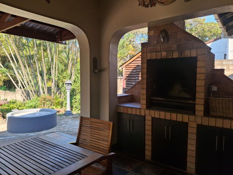 Covered patio area featuring a built-in brick braai, tiled flooring, and arched openings overlooking the garden and outdoor pool area.