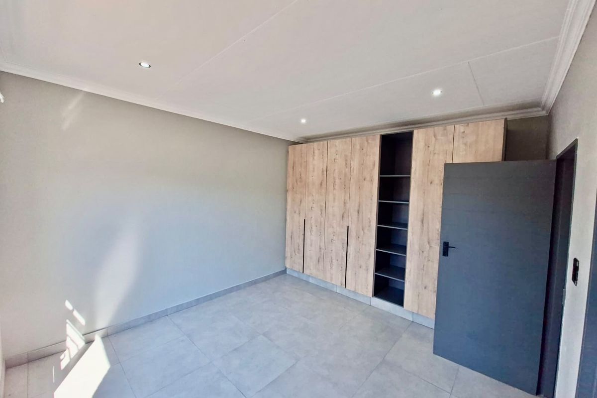 A bright, modern bedroom featuring neutral tiled flooring, recessed downlighting, and extensive built-in wooden cabinetry with open shelving.