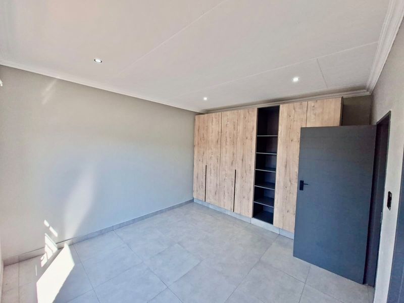 A bright, modern bedroom featuring neutral tiled flooring, recessed downlighting, and extensive built-in wooden cabinetry with open shelving.