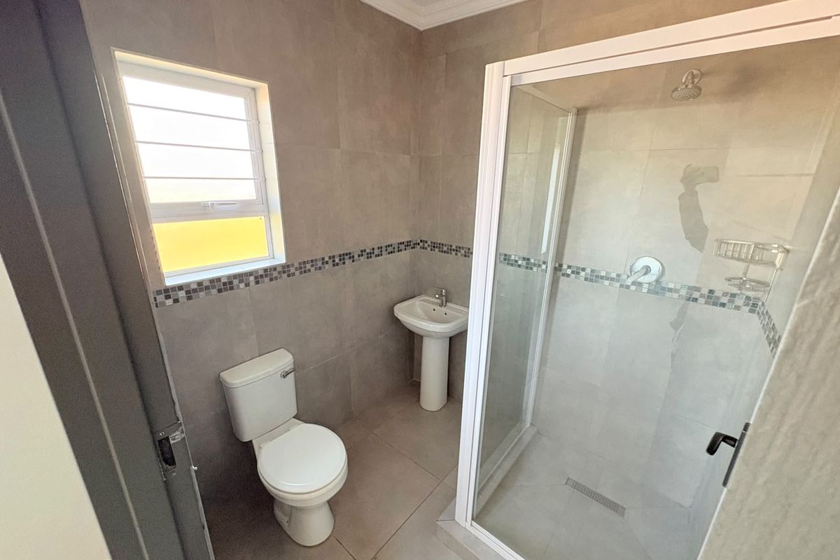 A clean, modern bathroom featuring a glass-enclosed shower, pedestal basin, and neutral floor-to-ceiling tiling with a decorative mosaic border.