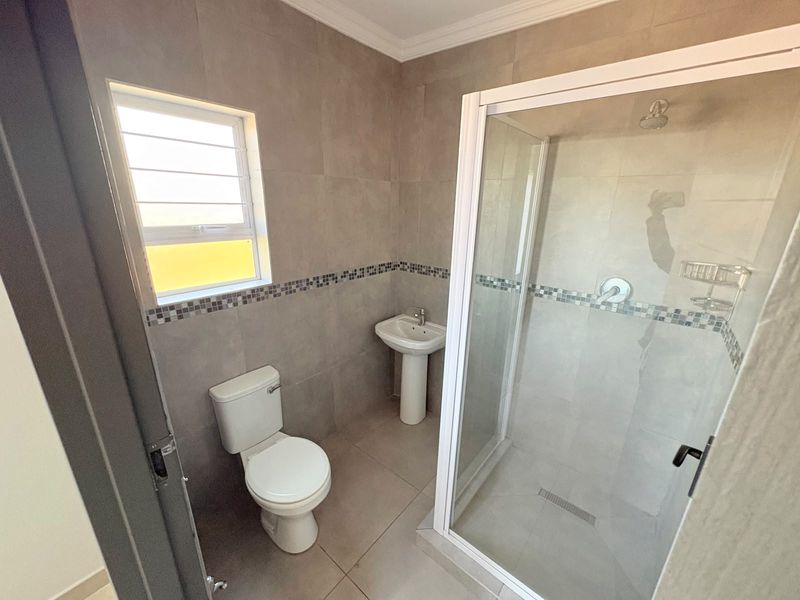 A clean, modern bathroom featuring a glass-enclosed shower, pedestal basin, and neutral floor-to-ceiling tiling with a decorative mosaic border.