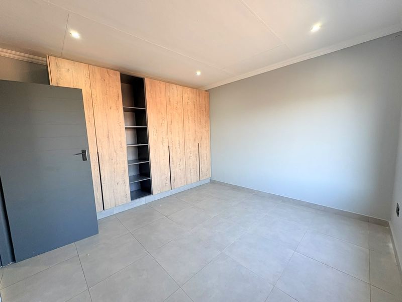A clean, empty bedroom featuring modern light wood-finish built-in cupboards, neutral tiled flooring, and recessed ceiling lighting.
