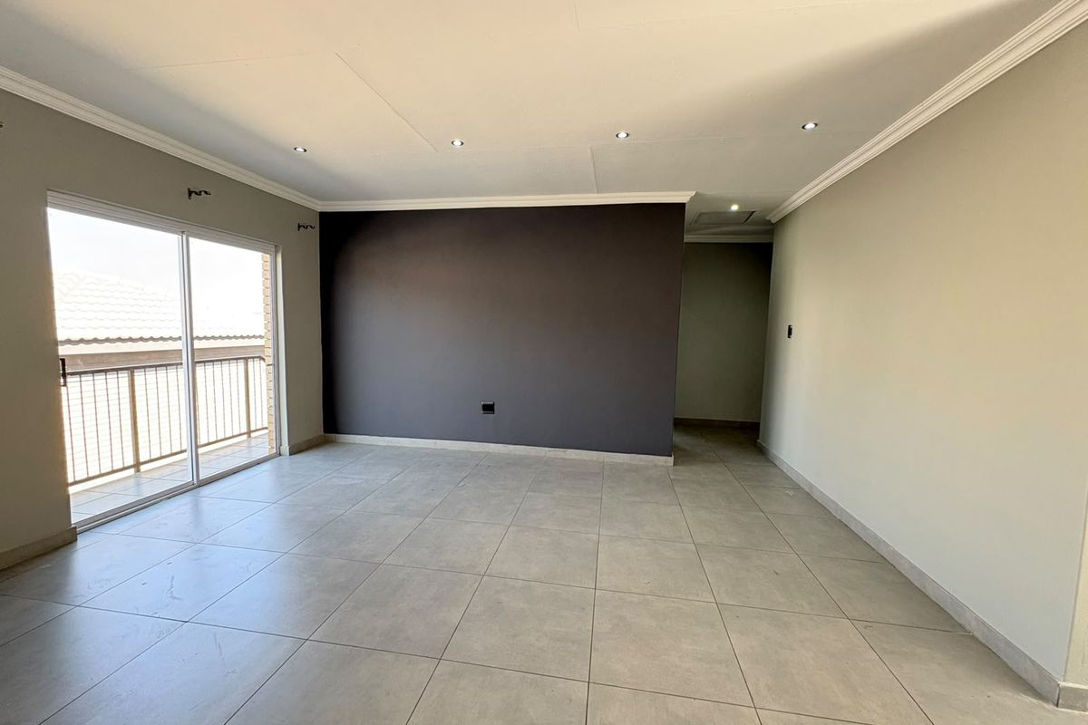 A spacious, empty living area featuring neutral tiled flooring, recessed downlighting, and a sliding glass door providing access to an outdoor balcony.