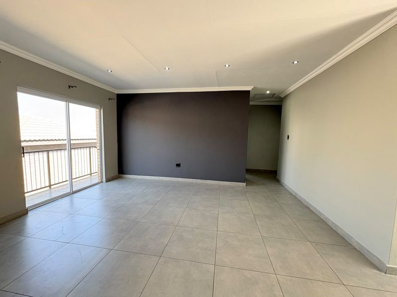 A spacious, empty living area featuring neutral tiled flooring, recessed downlighting, and a sliding glass door providing access to an outdoor balcony.