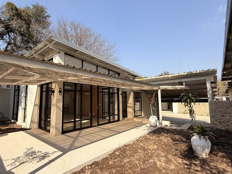 A modern rear exterior view featuring a covered patio, expansive black-framed glass doors, and clerestory windows, set against a landscaped garden area.