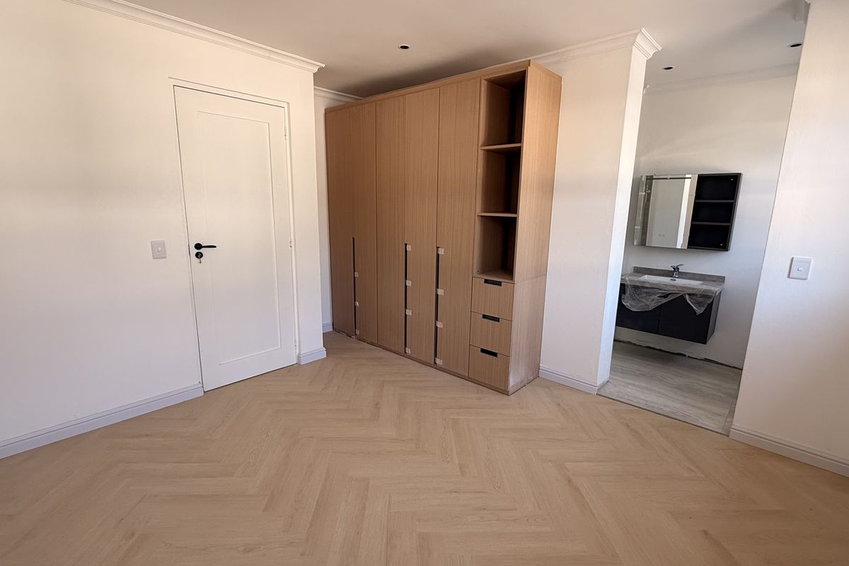 A bright, modern bedroom featuring light herringbone flooring, a large built-in wooden wardrobe, and an open-plan en-suite bathroom with a contemporary vanity.