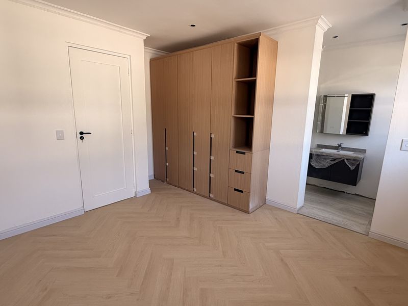 A bright, modern bedroom featuring light herringbone flooring, a large built-in wooden wardrobe, and an open-plan en-suite bathroom with a contemporary vanity.
