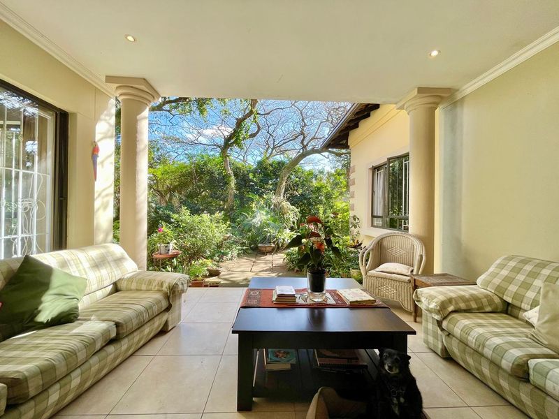 A spacious covered patio featuring tiled flooring, decorative columns, and recessed lighting, offering a seamless transition to the lush garden area.