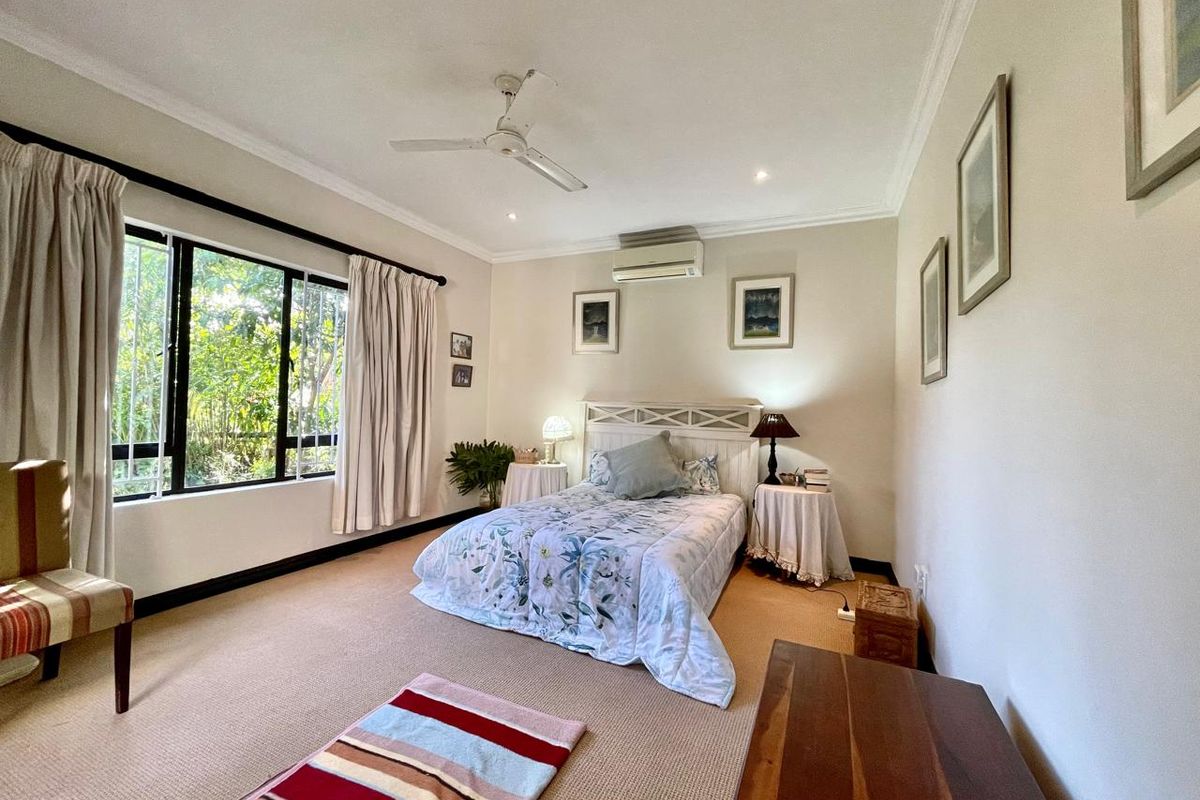 A spacious bedroom featuring neutral carpeting, a ceiling fan, and a wall-mounted air conditioning unit, with large windows providing natural light.