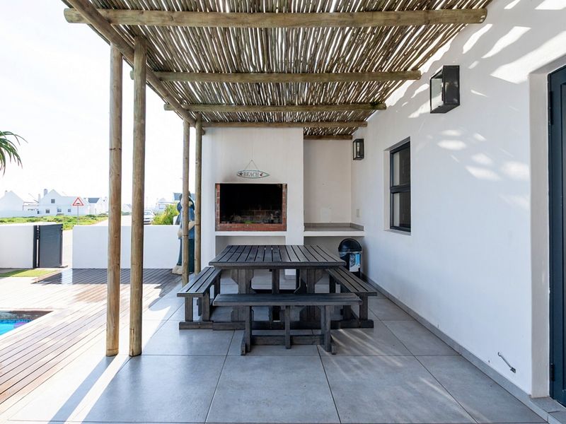 Covered outdoor patio featuring a built-in braai, timber pergola, and tiled flooring, overlooking a swimming pool and timber deck area.