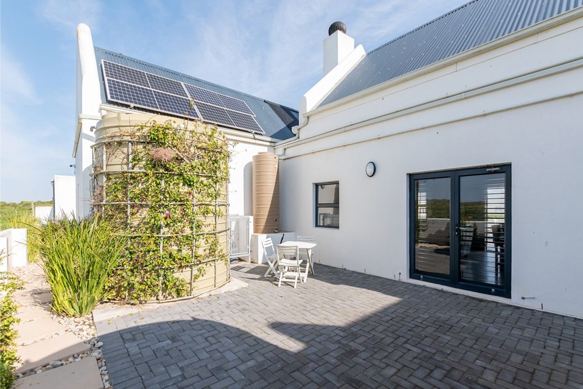 A paved rear patio area featuring a large water storage tank with climbing greenery, solar panels on the roof, and double glass door access.