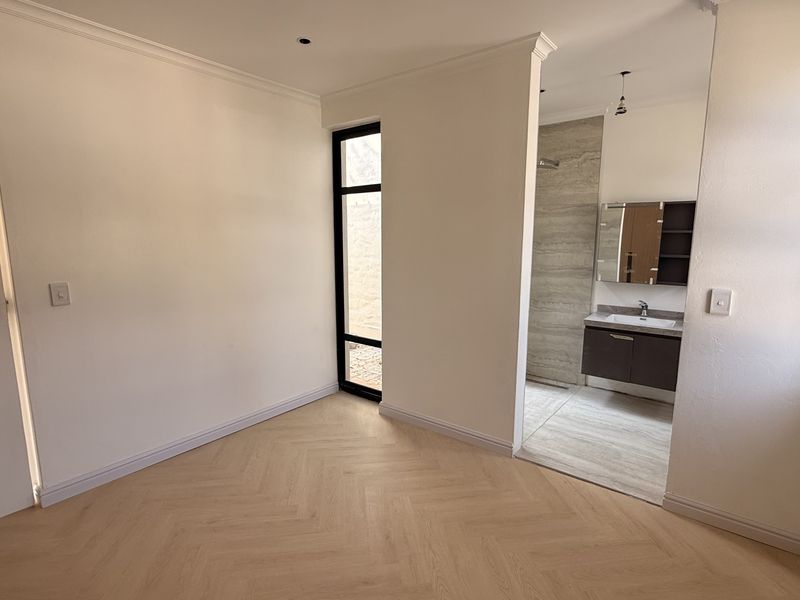 A bright, modern bedroom featuring light herringbone timber flooring, crisp white walls, and an open-plan en-suite bathroom with a contemporary vanity.