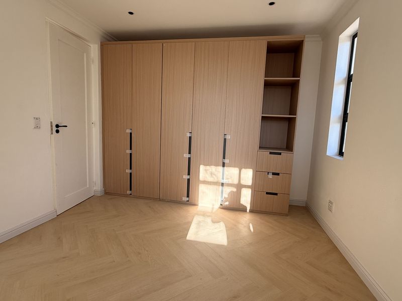 A bright, empty bedroom featuring modern light wood built-in wardrobes, herringbone timber flooring, and recessed downlights.