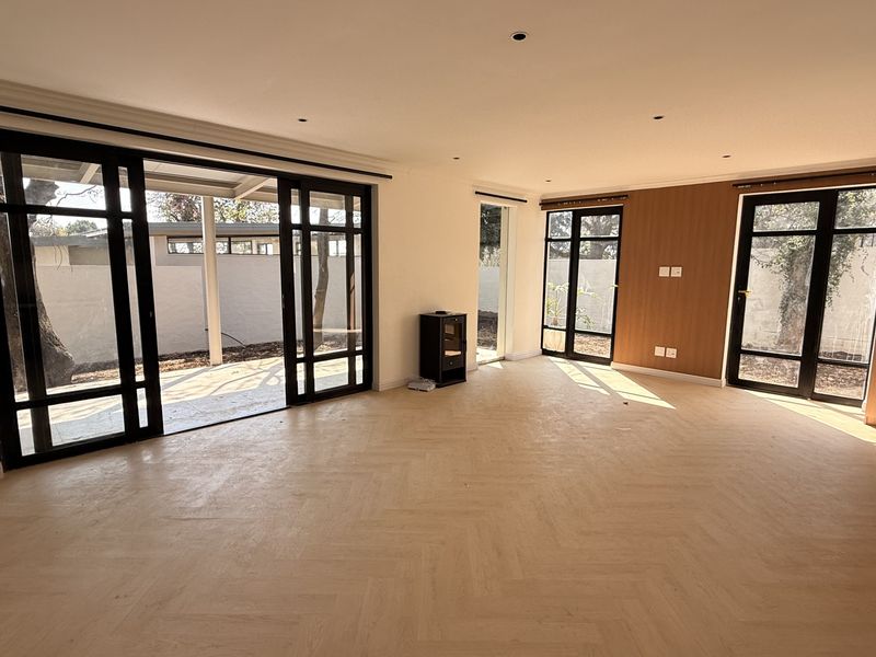 Spacious open-plan living area featuring light herringbone timber flooring, modern black-framed glass doors, and a cozy wood-burning fireplace with integrated kitchen cabinetry.