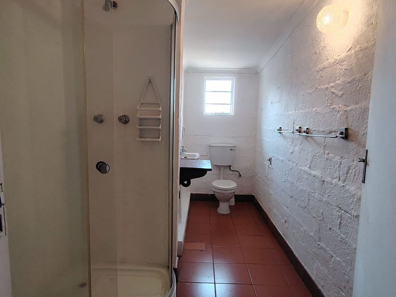 A functional bathroom featuring a curved shower enclosure, wall-mounted basin, and toilet, finished with terracotta floor tiles and textured white walls.