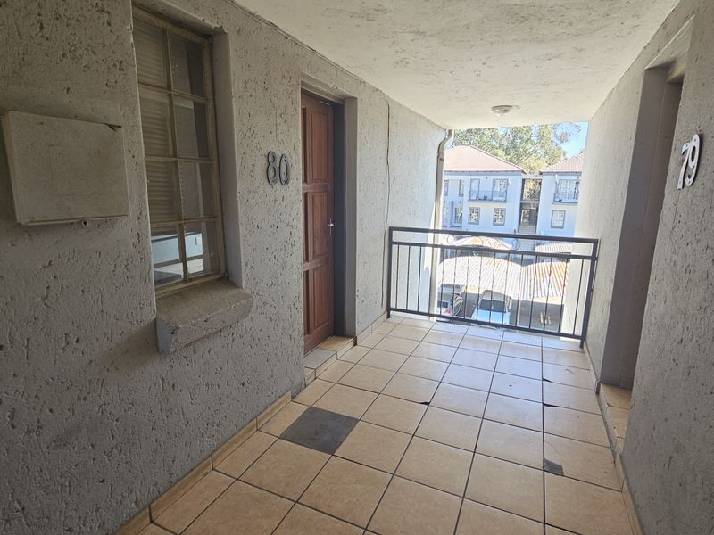 An exterior apartment balcony featuring tiled flooring, a metal balustrade, and a timber entrance door, showing signs of wear and tile damage.