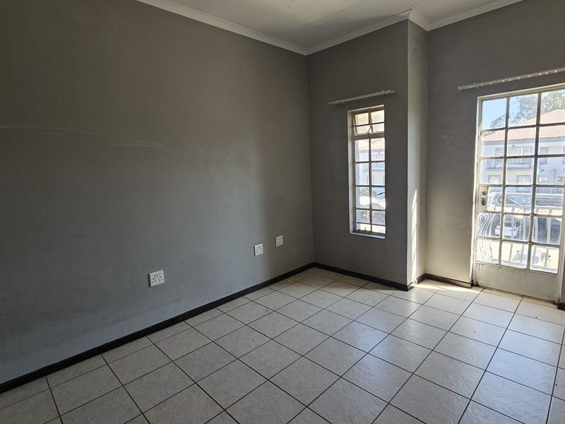 An empty living room featuring neutral grey walls, ceramic floor tiles, and natural light provided by a glass-paneled door and window.