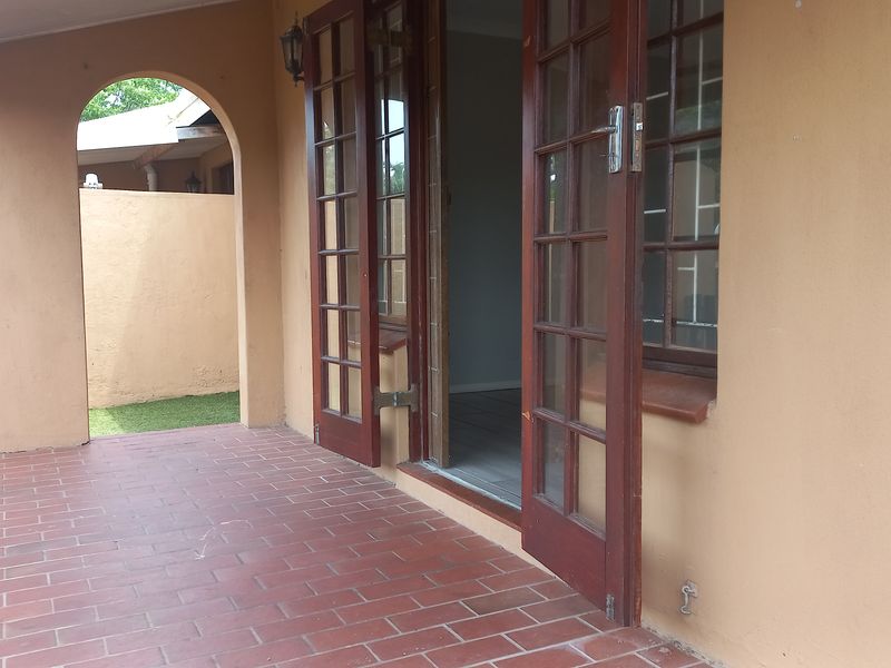 A covered patio area featuring red brick flooring, an arched architectural detail, and wooden french doors providing access to the interior living space.