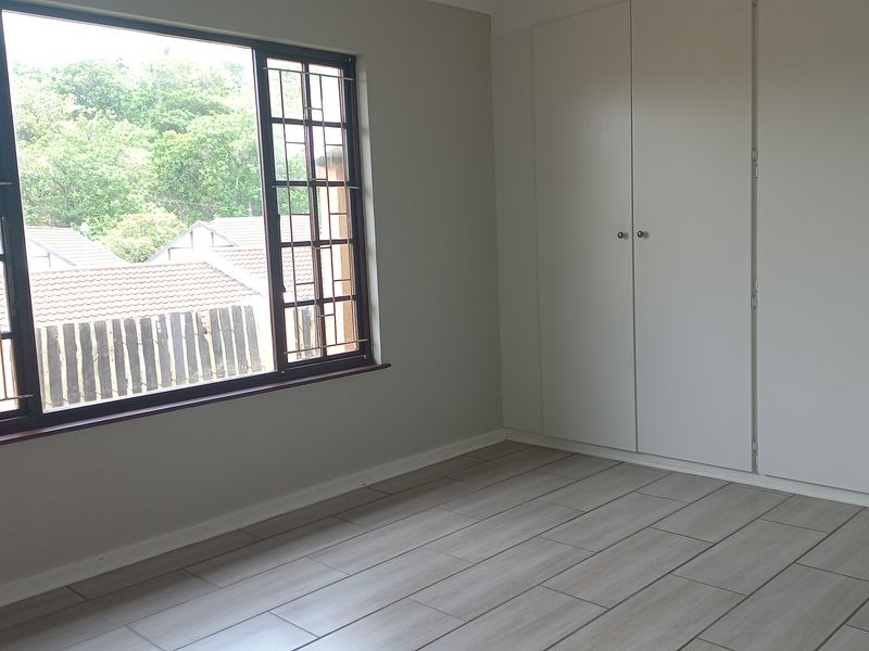 A bright, empty bedroom featuring neutral tiled flooring, built-in cupboards, and a large window with security bars overlooking the neighborhood.