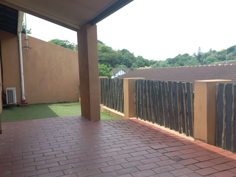 A covered patio area featuring brick paving, artificial grass, and a timber slat balustrade overlooking the surrounding neighborhood.
