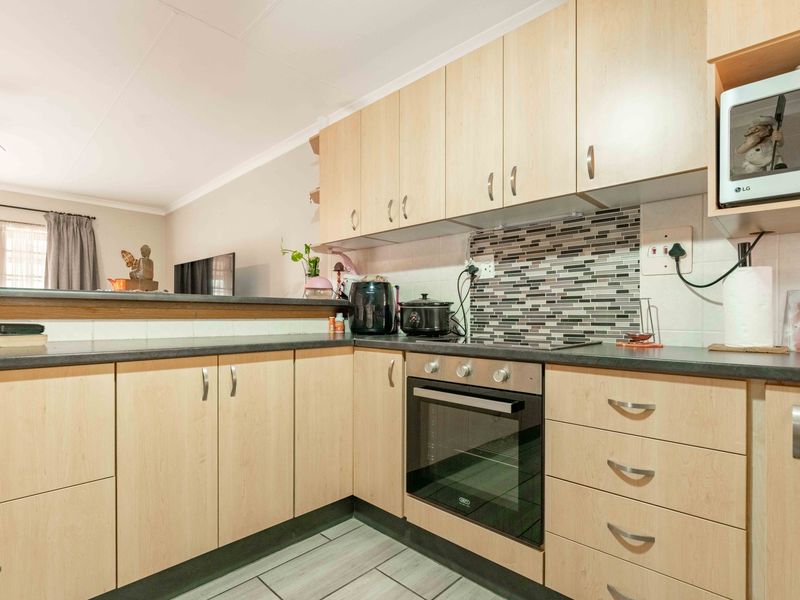 A functional kitchen featuring light wood-finish cabinetry, dark countertops, a mosaic tile backsplash, and integrated oven and hob, opening into the living area.