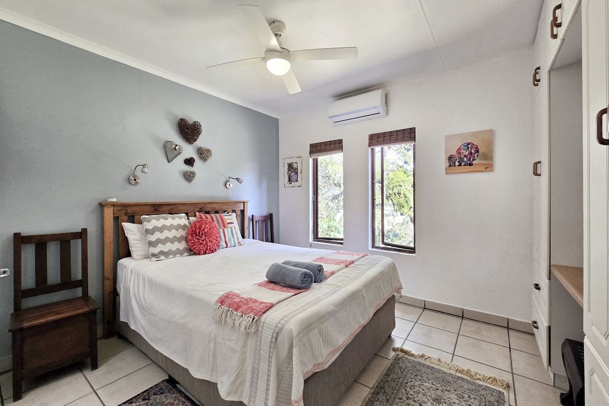 A well-lit bedroom featuring tiled flooring, built-in cupboards, a ceiling fan, and a wall-mounted air conditioning unit.