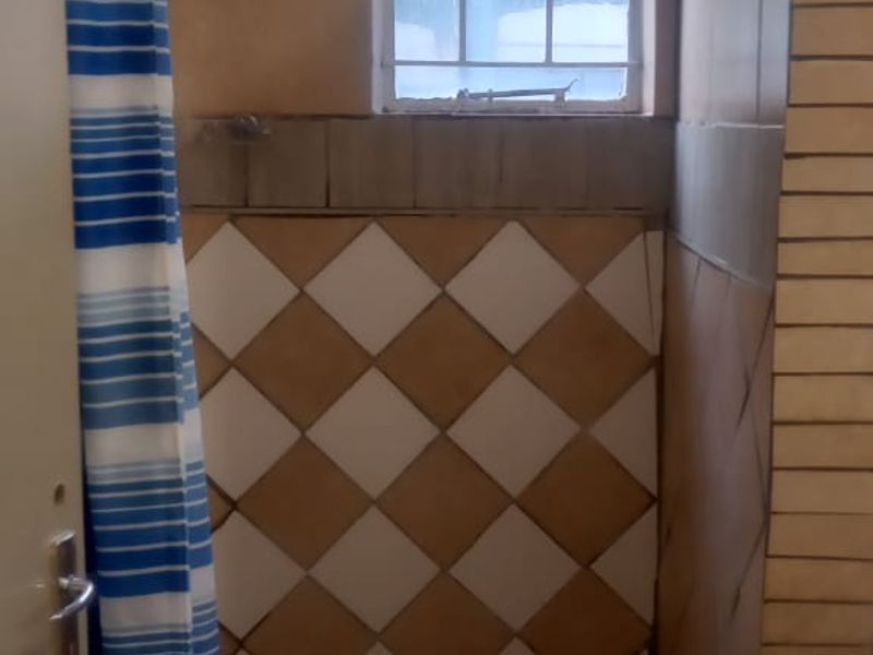 A dated shower enclosure featuring tiled walls and flooring, equipped with a fixed shower head and a small window with security bars.