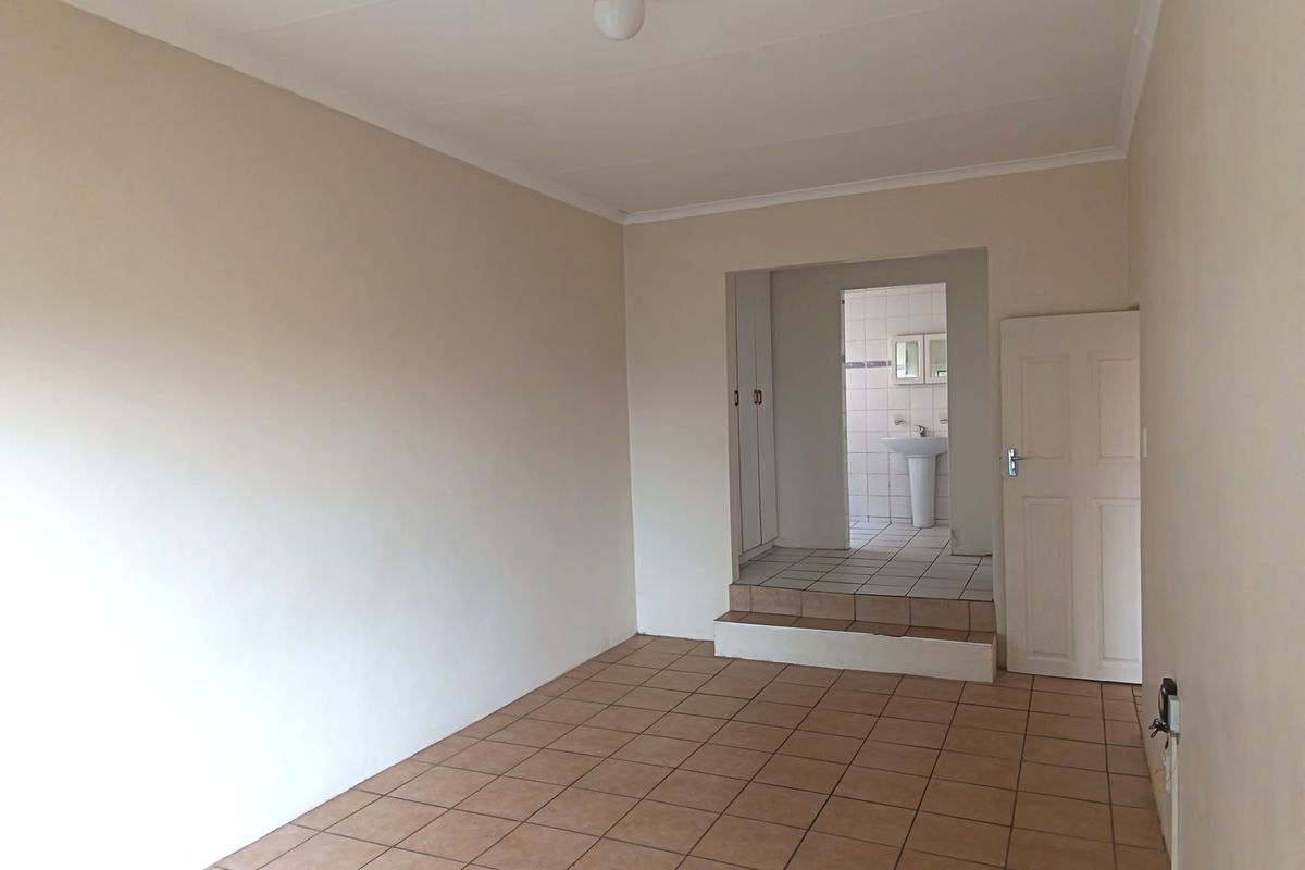 An empty bedroom featuring tiled flooring, built-in cupboards, and an adjoining en-suite area with a pedestal basin.