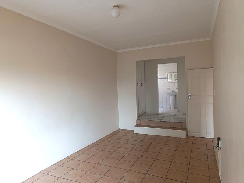 An empty bedroom featuring tiled flooring, built-in cupboards, and an adjoining en-suite area with a pedestal basin.